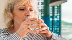 Elderly woman in a striped shirt drinks water with a person comforting her with a hand on her shoulder in a medbridge setting.