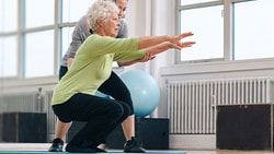 Older adult performing a balance squat with support from a physical therapist in a gym setting for fall prevention training.