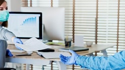 Masked and gloved individuals exchange a document in an office with computers and large windows.