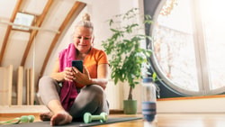 Older woman on a yoga mat smiles at her phone while using medbridge, with dumbbells and a water bottle nearby.