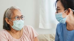 An elderly woman and a nurse in a blue uniform, both masked, talk indoors.