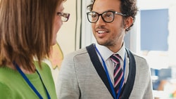 Two professionals with name badges converse near a wall of maps, both wearing glasses.
