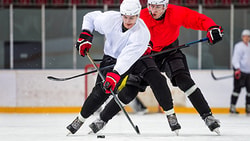 Two ice hockey players, one in white and one in red, compete for the puck as two others are visible in the background on an ice rink.