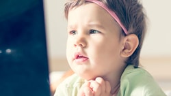 Toddler with a pink headband watches a screen, chin on hands.