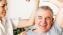 A Medbridge therapist helps an elderly man with arm exercises in a bright room.