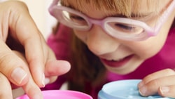 A child with glasses happily plays with containers, helped by an adult hand.