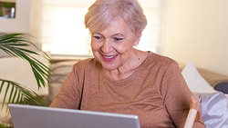 Smiling older woman using a laptop on a couch, surrounded by indoor plants.