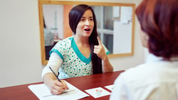 A woman in scrubs gestures while speaking to another woman at a Medbridge office desk.
