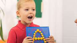 A child holds a blue board with yellow pegs forming "A," watched by an adult, with a plant in the background.
