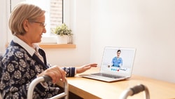 An elderly woman in a wheelchair video calls a healthcare professional on a medbridge laptop.