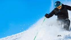 Skier in black gear with green poles on a snowy slope beneath a blue sky.
