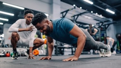 A man does push-ups in a gym as another observes, with medbridge digital care solutions implied for fitness support.