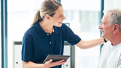 Female clinician smiling and holding a tablet while talking to an older male patient during a check-in, representing patient engagement.