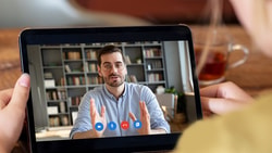 A video call on a tablet shows a man speaking, with bookshelves in the background, beside a tea cup and phone on the table.