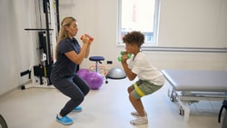 A physical therapist and a boy perform squats with dumbbells while using Medbridge patient care software.