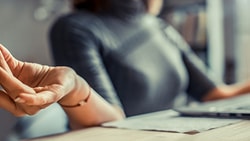 Person meditating at a desk while using a laptop with a coffee cup nearby, representing Medbridge balance.