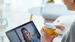 Woman sits on a couch holding a coffee mug while speaking to a healthcare provider via telehealth video call on a laptop.