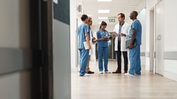Group of clinicians in scrubs and a lab coat conferring over paperwork in a hospital corridor