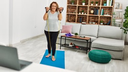 A woman exercises on a blue yoga mat in a bright living room with a laptop and furniture in the background.