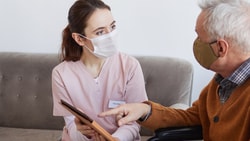 A nurse in pink and a mask holds a tablet, talking to an elderly masked man on a sofa at home.