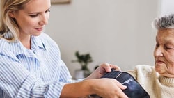 In a bright living room, a caregiver helps an elderly woman use a blood pressure monitor.