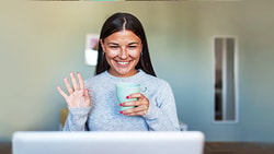 A person smiles, holding a mug and waving at a laptop on a Medbridge video call.