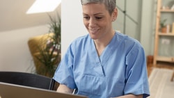 A person in blue scrubs works on a laptop at a desk, with Medbridge materials visible.