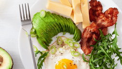 A breakfast plate with avocado, egg, bacon, arugula, cheese, plus an avocado half and juice nearby.