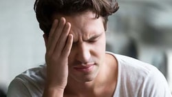 A person in a white shirt appears distressed with their hand on their forehead, background blurred.