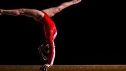 A gymnast in a red leotard doing a handstand on a balance beam.