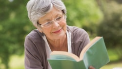 Elderly woman with glasses reading a green book outside amid lush greenery.