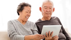 An elderly couple smiles while looking at a tablet on a couch, with a softly lit background.