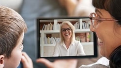 A child and an adult have a video call on a tablet with a smiling woman in glasses in front of a bookshelf, linked to medbridge.