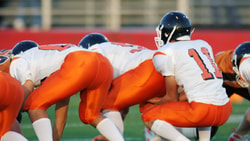Football players in white and orange uniforms are lined up at the start of a game on Medbridge field.