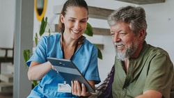 A healthcare worker and an older man smile while looking at a tablet in a bright room.