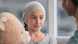 A young child with a headscarf holds a teddy bear, sitting opposite a woman in a medbridge uniform.
