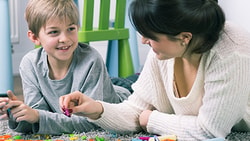 A woman and child build with blocks on a rug, with colorful chairs behind them.