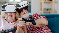Elderly couple using VR headsets and controllers, smiling on a couch.