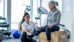 A Medbridge therapist helps an elderly woman with leg exercises in a gym.