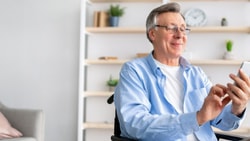 A smiling older man in a wheelchair uses a smartphone, with books and plants in the background.