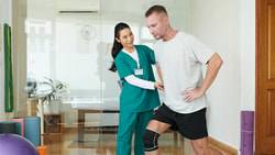 A physical therapist helps a man in a knee brace with balance exercises in a Medbridge therapy room.