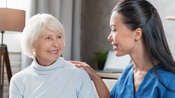 An elderly woman with white hair smiles at a younger woman in blue scrubs who is touching her shoulder.