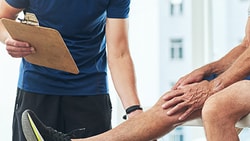 A man in athletic wear uses a clipboard to assess a seated patient's knee at medbridge.