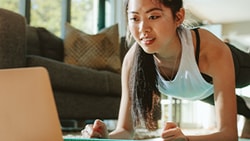 A woman does a plank exercise indoors while watching a medbridge video on a laptop.