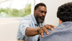 A bearded man offers comfort to someone beside a large window and beige couch in the medbridge room.