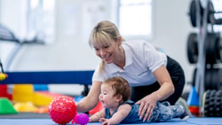 A woman uses medbridge digital healthcare to help a baby on a mat reach for red and purple toys in a therapy session.