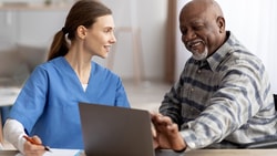 A Medbridge nurse in blue scrubs discusses a laptop screen with a man in a wheelchair.