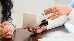 A person in a white sweater and another in scrubs discuss a medbridge pamphlet at a table with a folder.