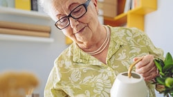 Elderly woman with glasses and floral shirt watering indoor plants.