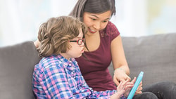 A woman points at a tablet while sitting on a couch with a child in glasses and a plaid shirt.